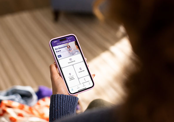 A hand holds a phone displaying the MyCHRISTUS app. The screen displays On Demand Care, MyCHRISTUS , Maps & Locations and Virtual visits.